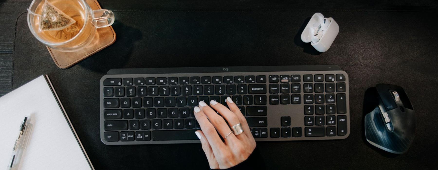 woman's hand on a keyboard surrounded by office items and a cup of tea on a coaster